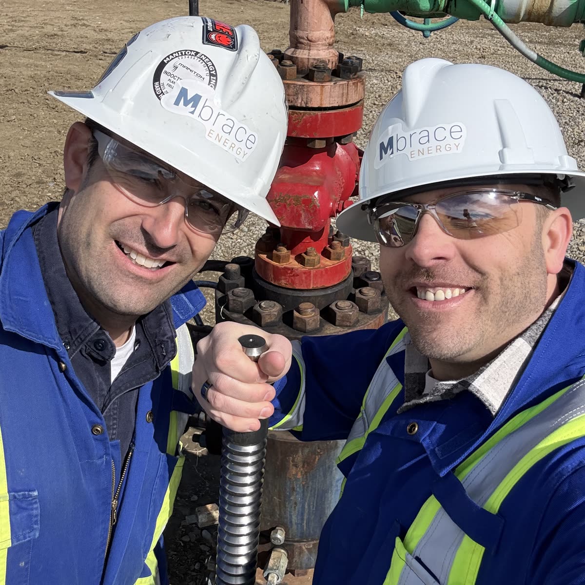 Joshua Anhalt and Steven Tulissi in Mbrace Energy hard hats at a wellhead, holding the well's plunger